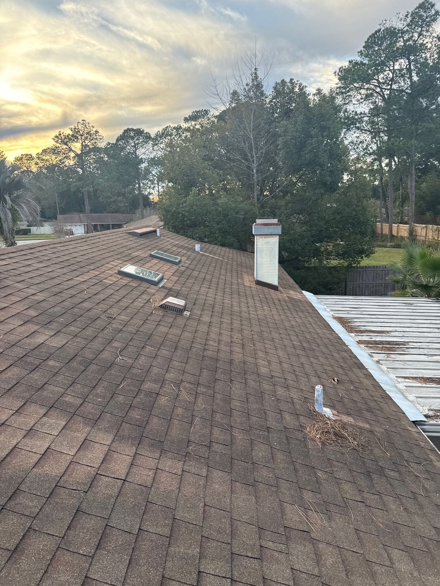 Brown shingle roof with vents and chimney against a cloudy sky, trees, and partial view of a white metal roof.