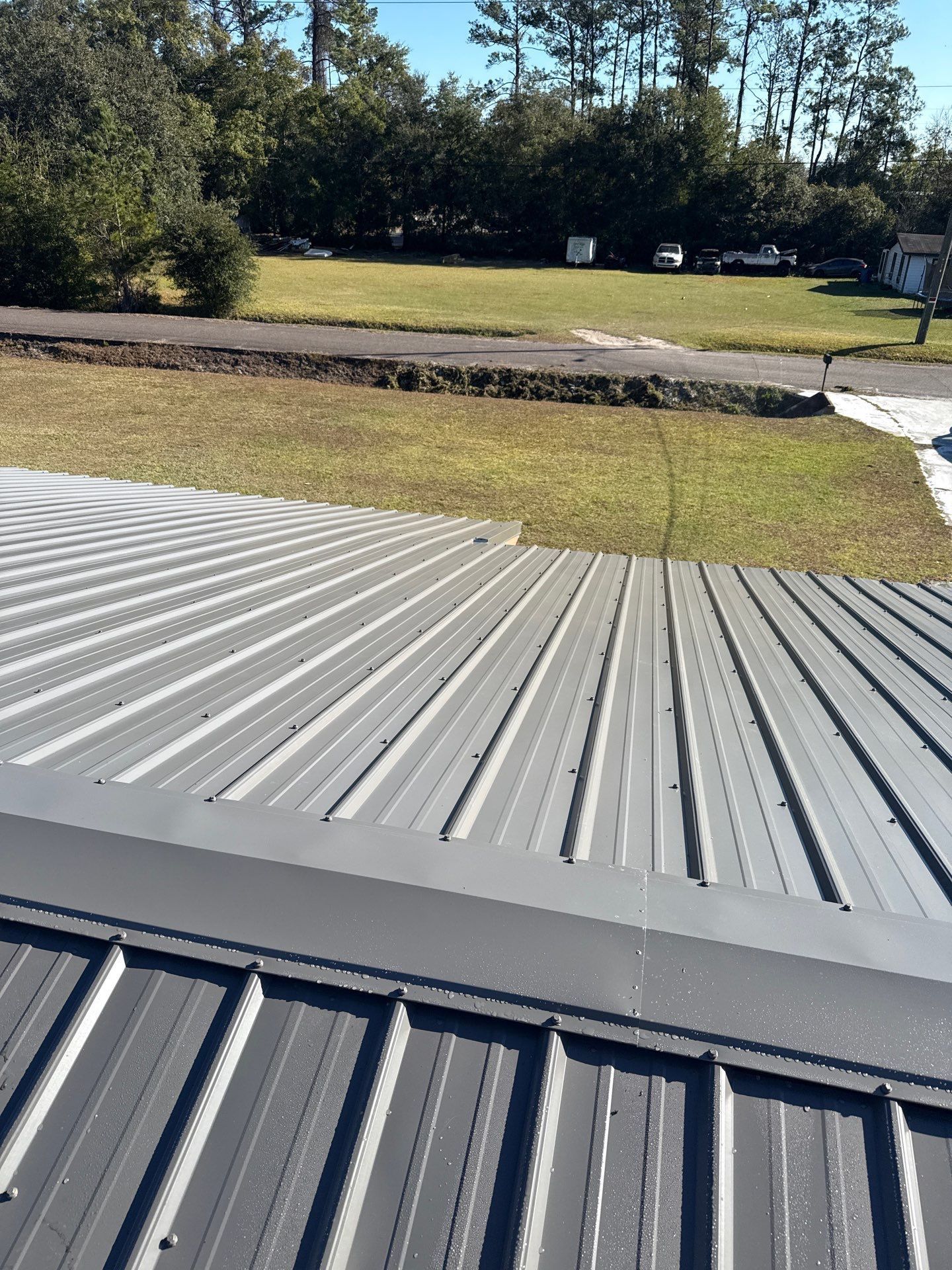 Gray metal roof with ridges, angled toward a yard with grass and trees under a clear blue sky.