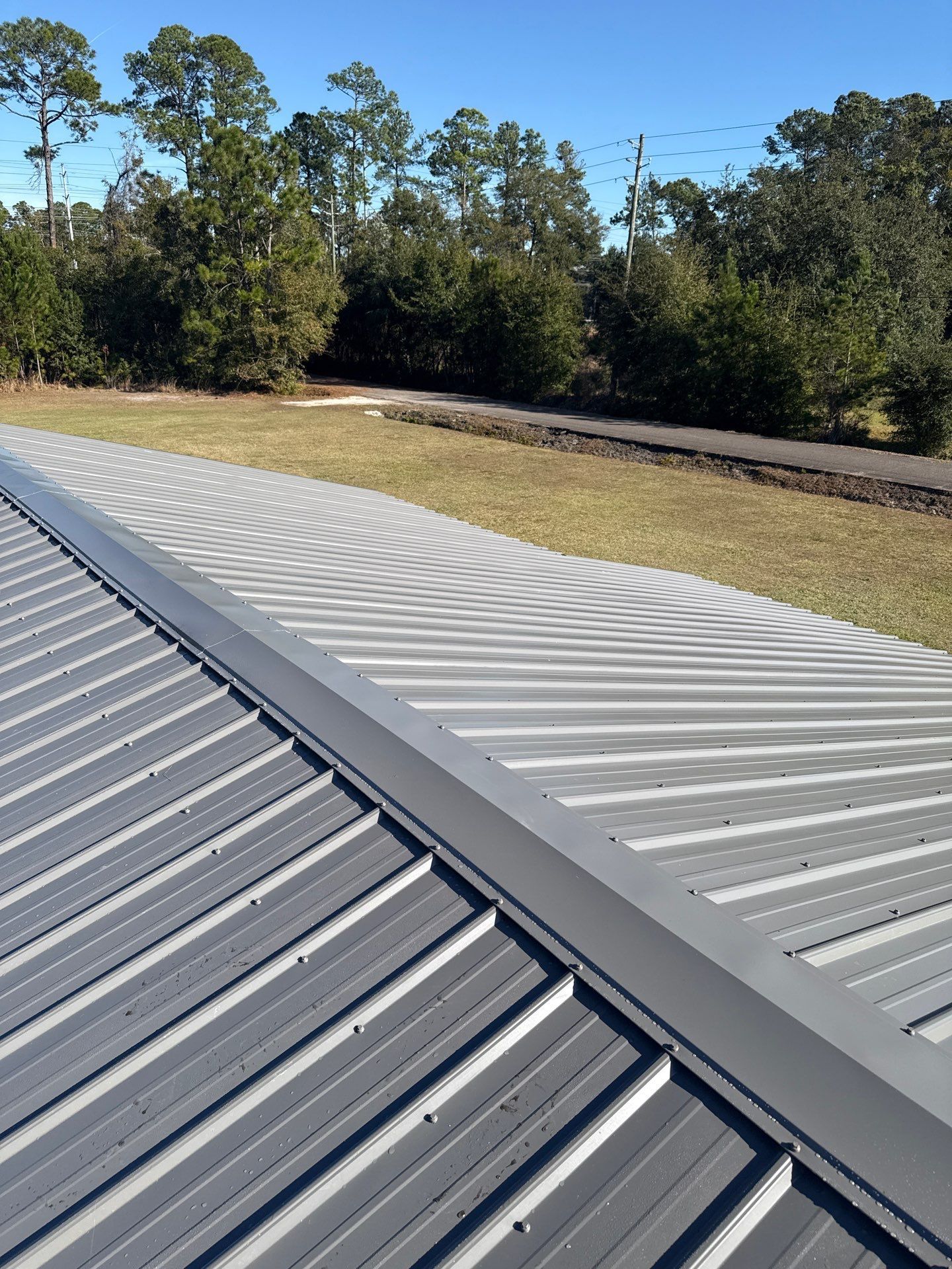 Close-up of a gray metal roof with ridged panels. The roofline has a flat section along the ridge. Sunny outdoor setting.
