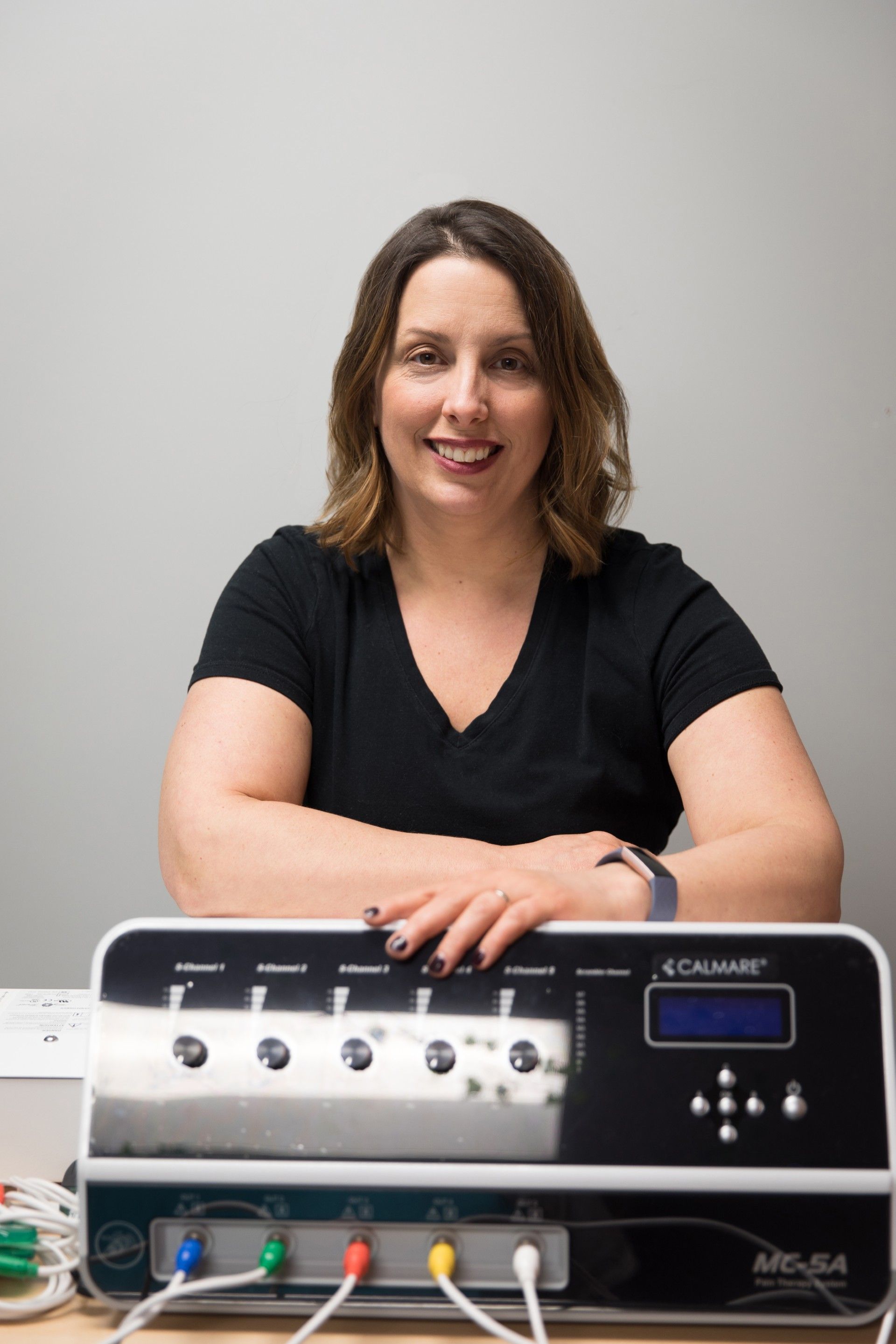 Woman smiling, leaning on a medical device with dials and a screen; gray wall background.