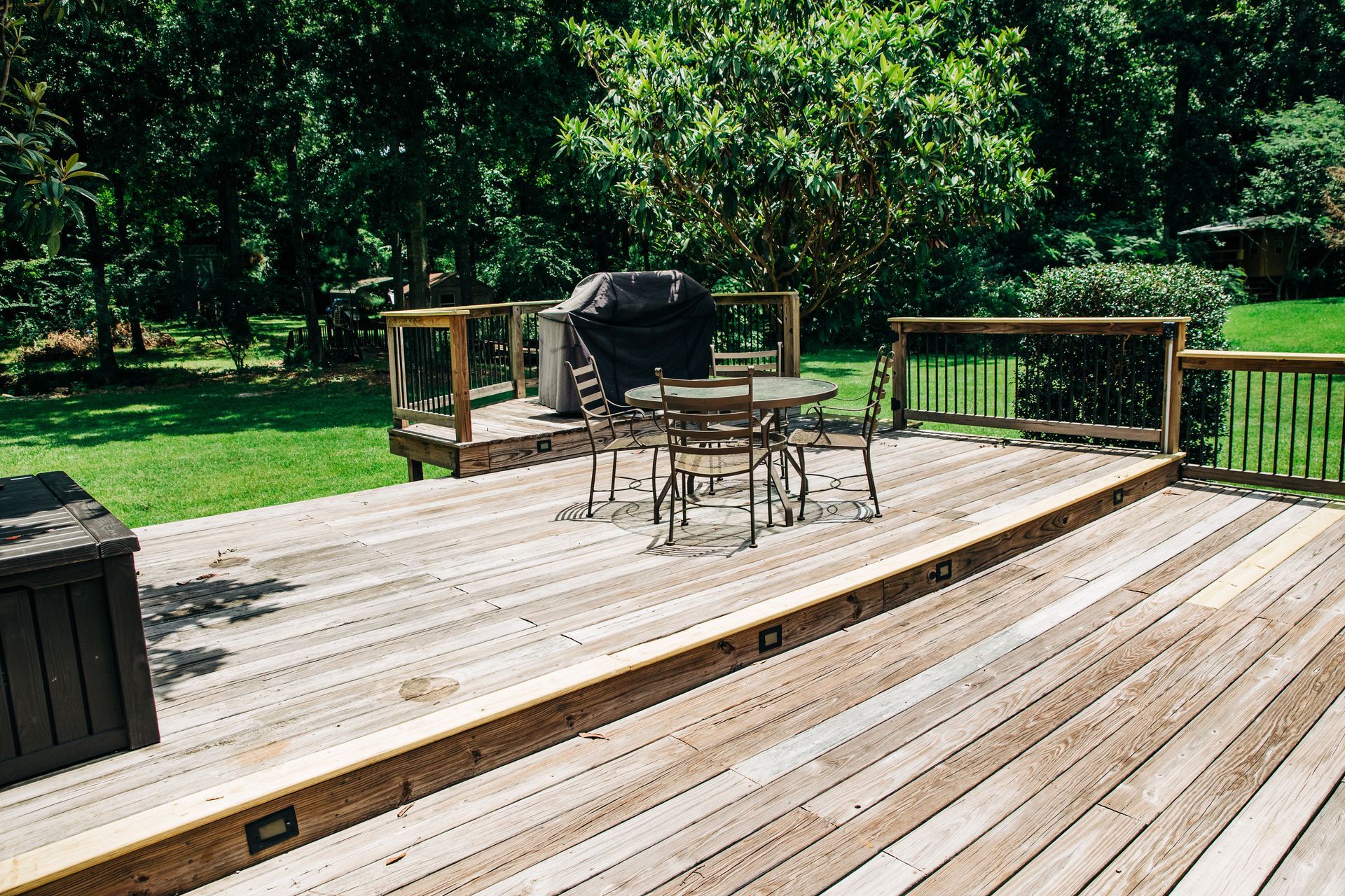 Wooden deck with outdoor table, chairs, grill, and railing overlooking a green yard with trees Wooden deck with outdoor table, chairs, grill, and railing overlooking a green yard with trees