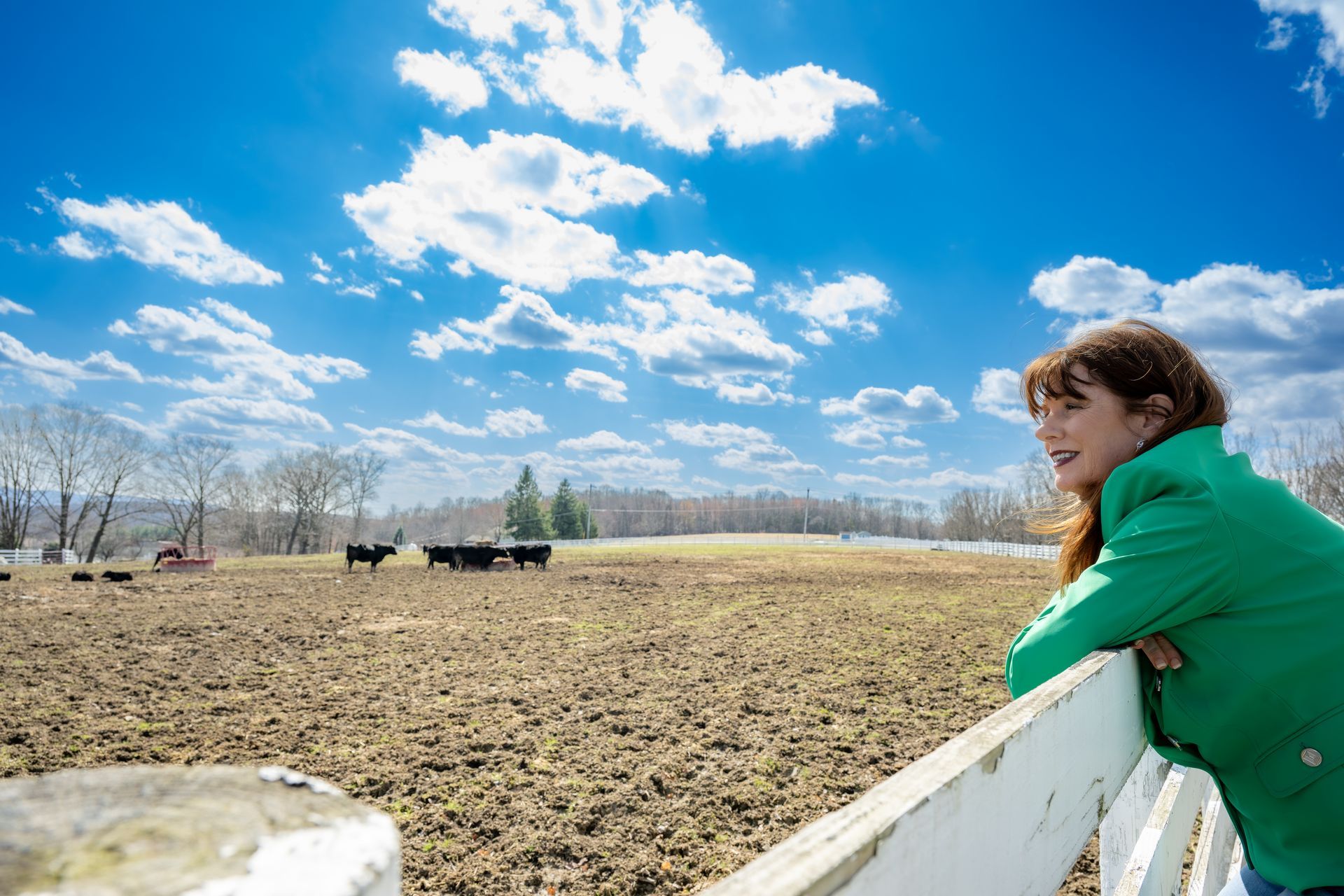 Woman leans on a white fence, smiles at a field with cows, under a blue sky with clouds.