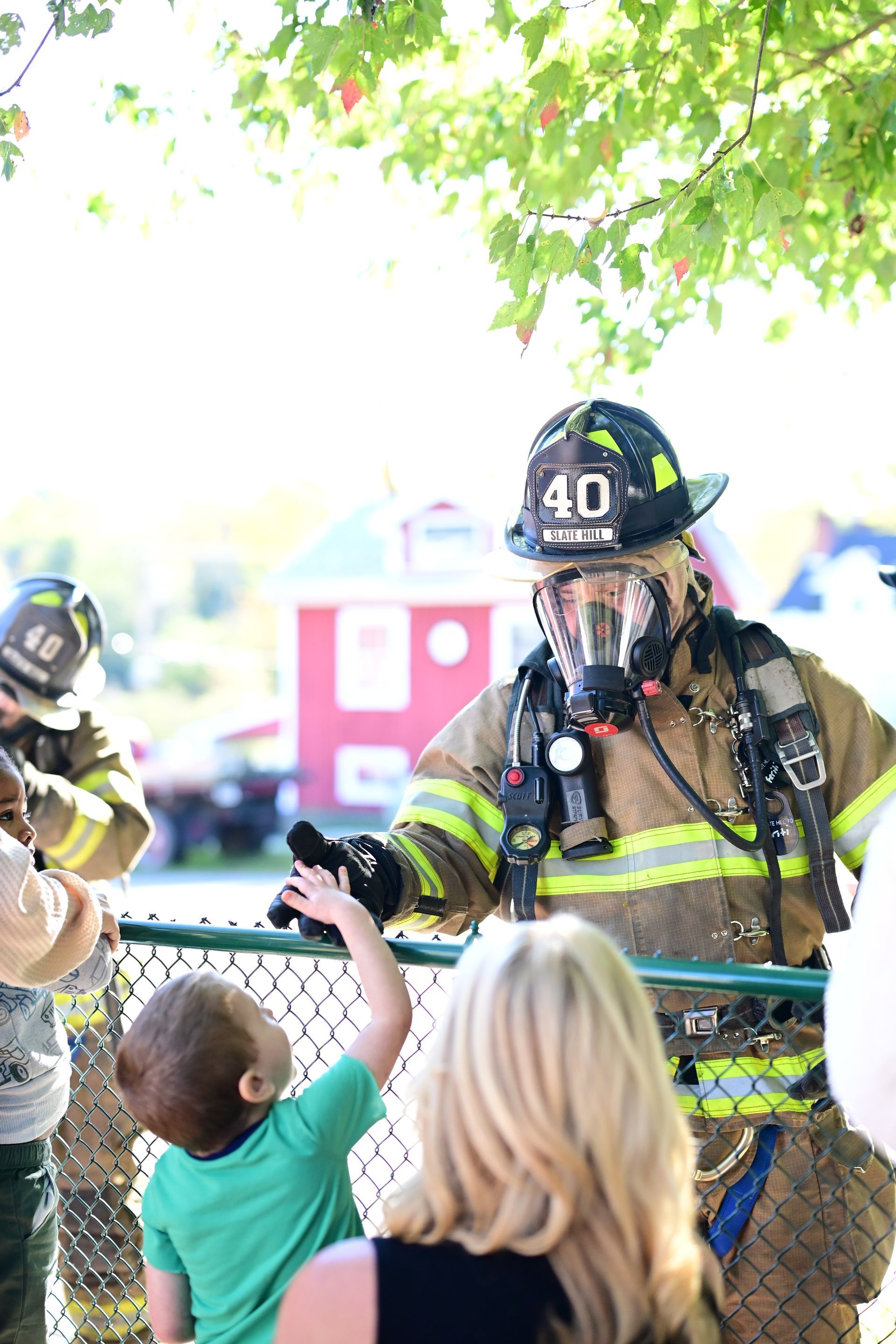 Firefighter in full gear interacting with a child reaching toward him; woman watching, outdoor setting.