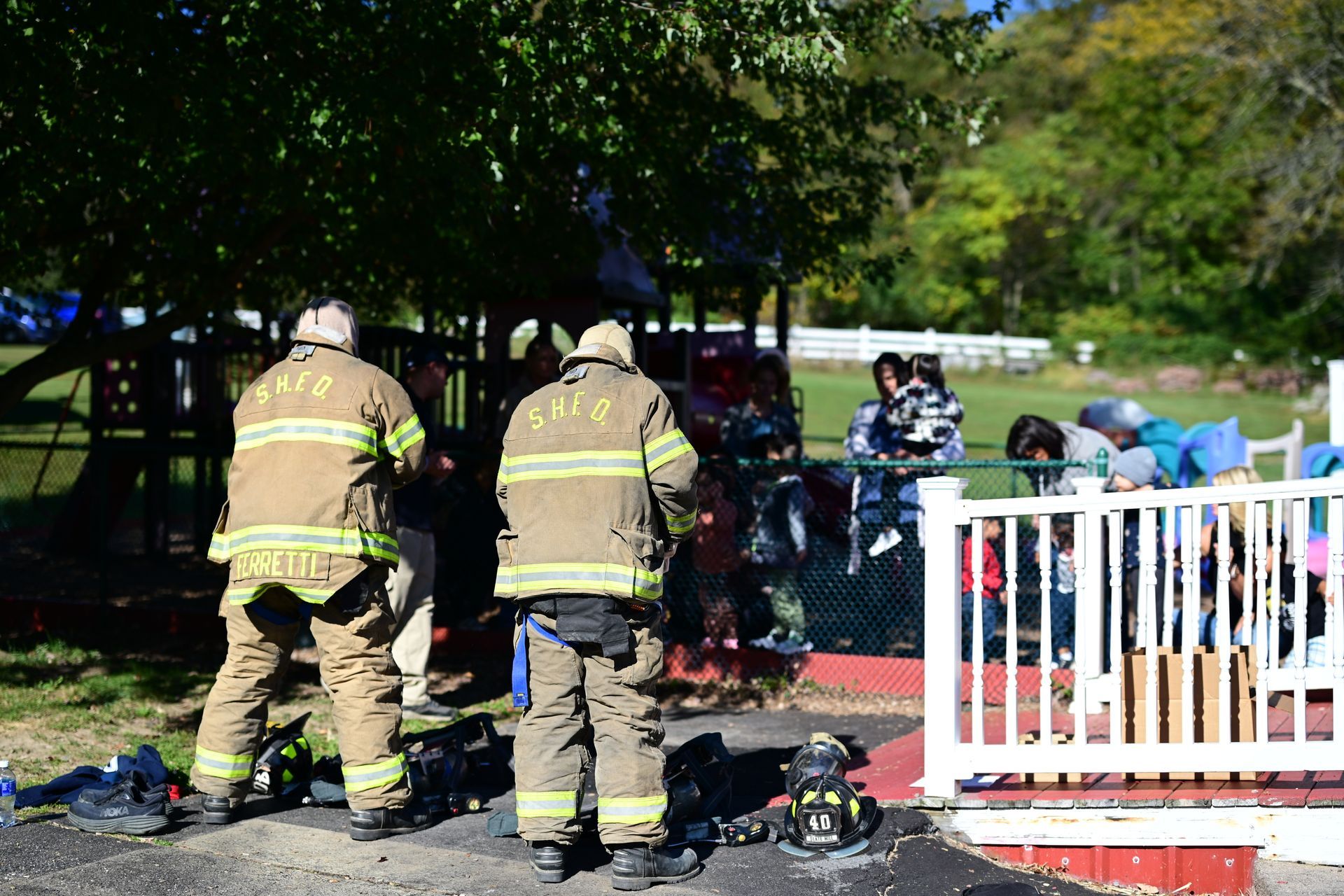 Firefighters at a playground inspect damage after a fire. Children watch from a fence.