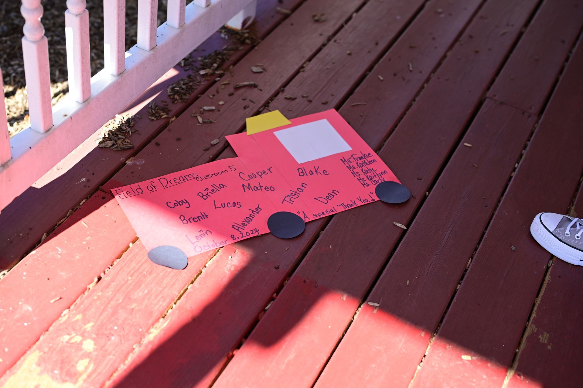 Red cardstock car on a wooden porch with black wheels and a white window; a person's foot is visible.