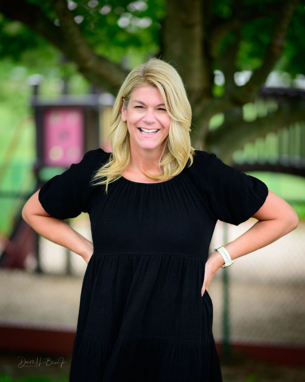 A woman in a black dress is standing in front of a playground with her hands on her hips.