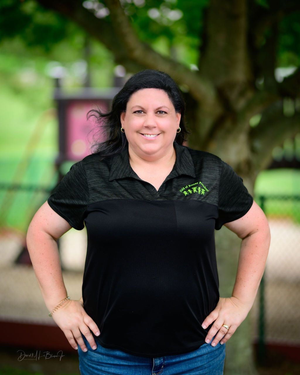 A woman in a black shirt is standing with her hands on her hips in front of a tree.