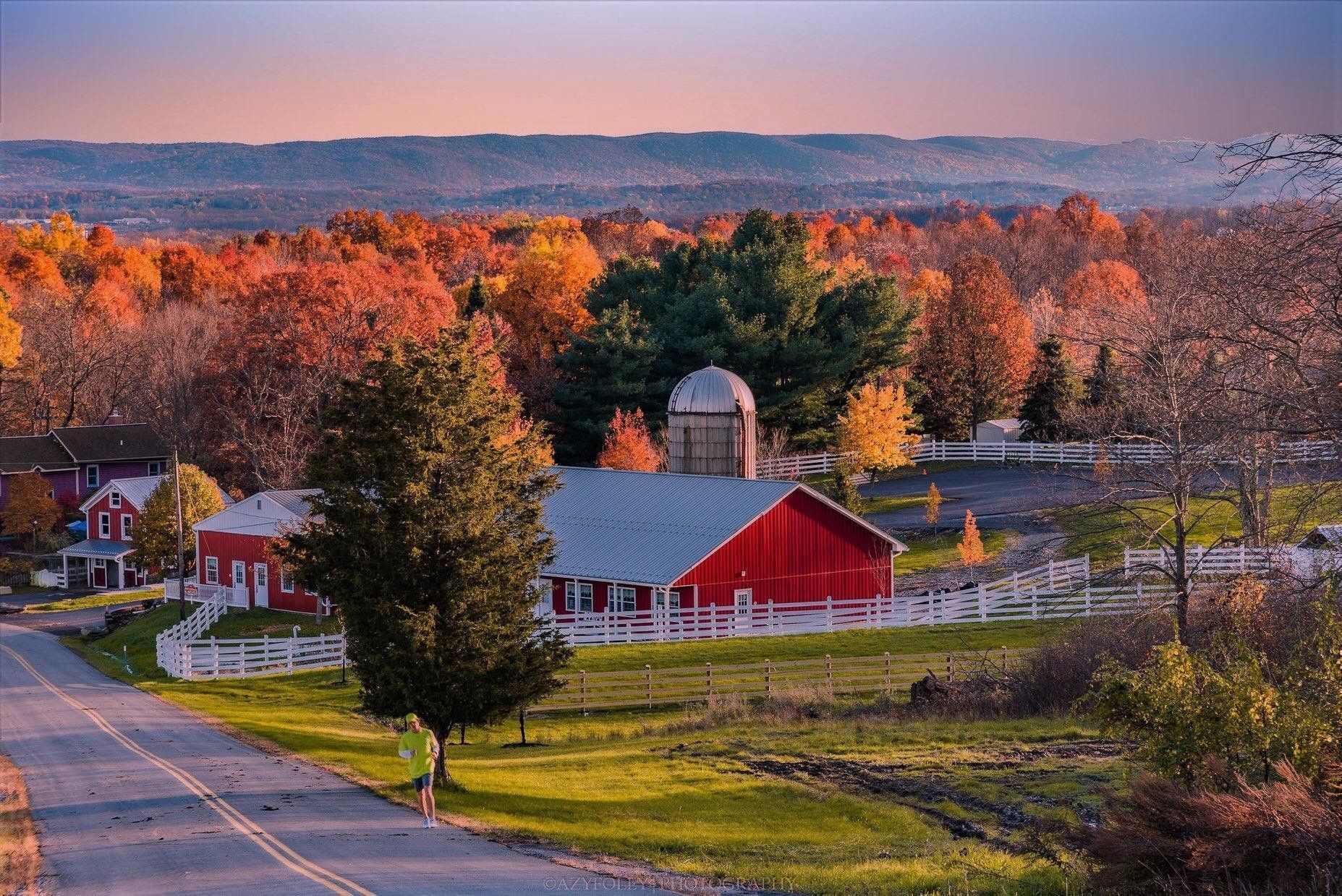 Red barn and silo in autumn landscape; road in foreground, colorful trees.