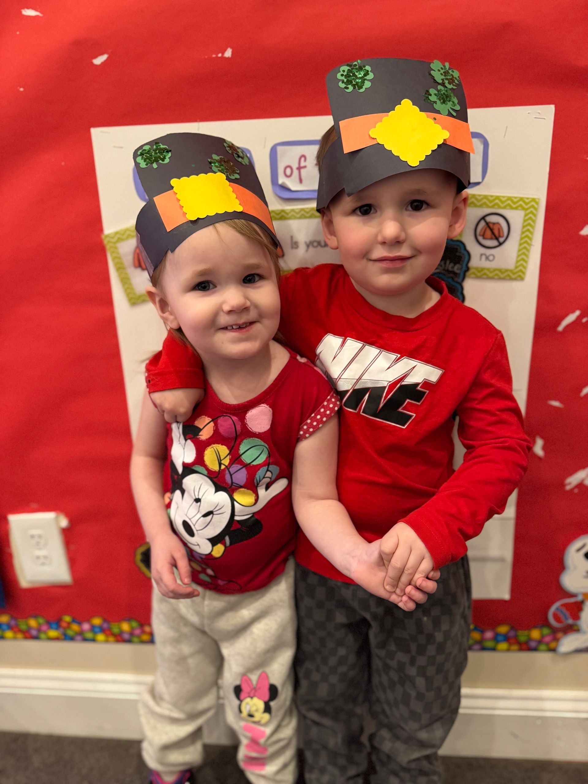 Two toddlers wearing pilgrim hats hug; red shirts, smiling in front of a poster.