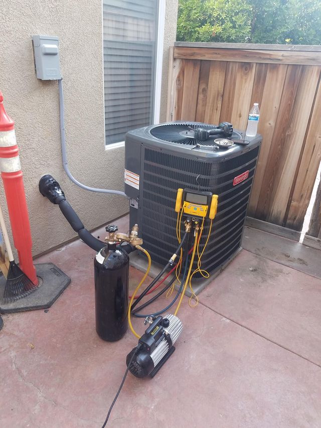 HVAC technician servicing a black air conditioning unit. Equipment includes gauges, vacuum pump, and refrigerant tank.