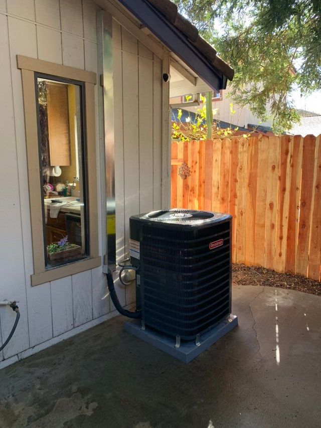 HVAC unit outside a building with a window and a silver vent pipe. Wooden fence in background.
