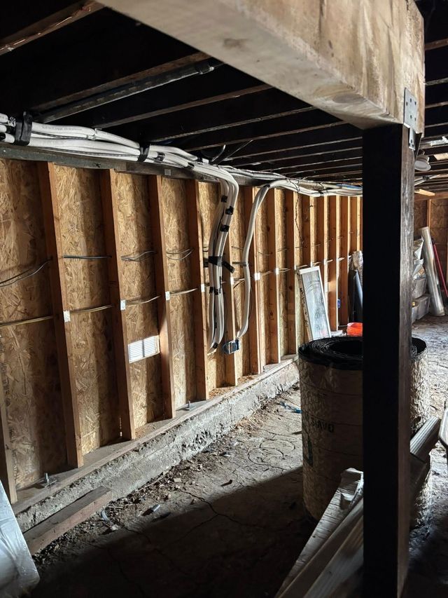 Wooden studs and beams are visible. Various wires and pipes hang from the ceiling and walls.