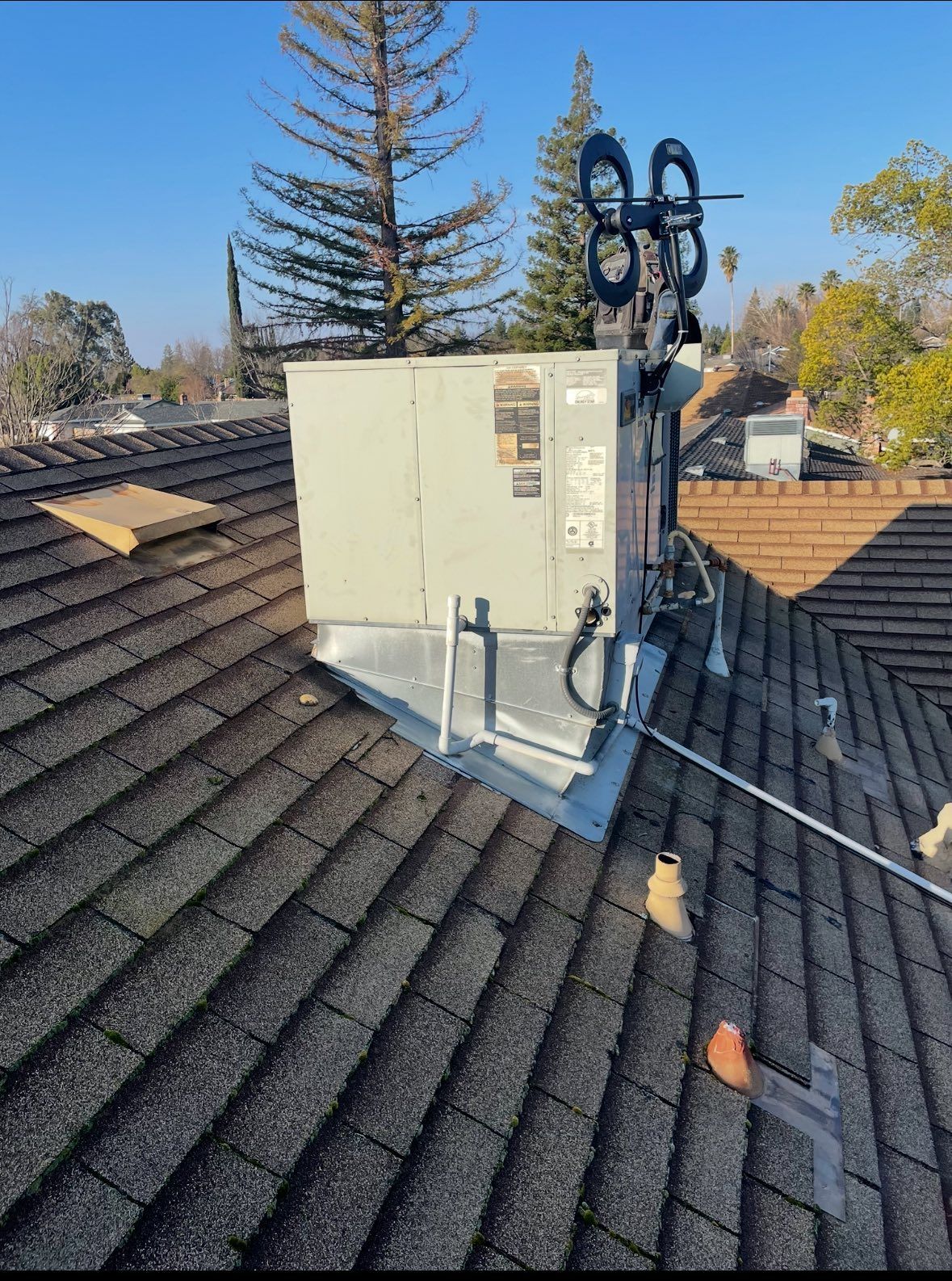 HVAC unit on a rooftop with a large antenna. Gray unit on a metal platform, shingles, and blue sky.