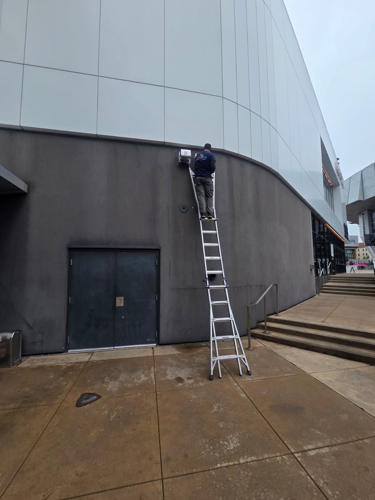 Person on a ladder, working on a building exterior. Gray walls, overcast sky.