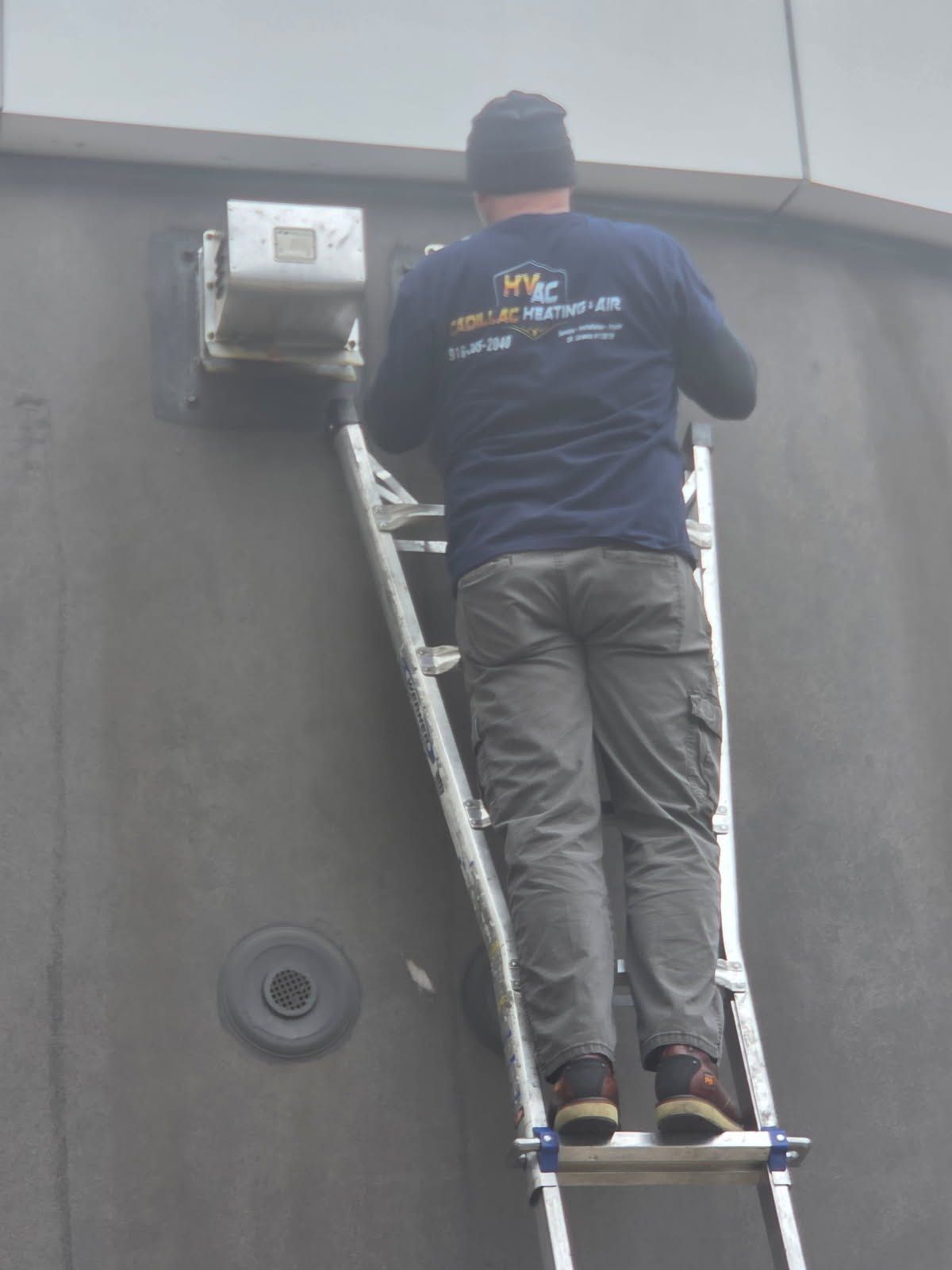 Man on a ladder, working on a gray building. He wears a blue shirt and gray pants.