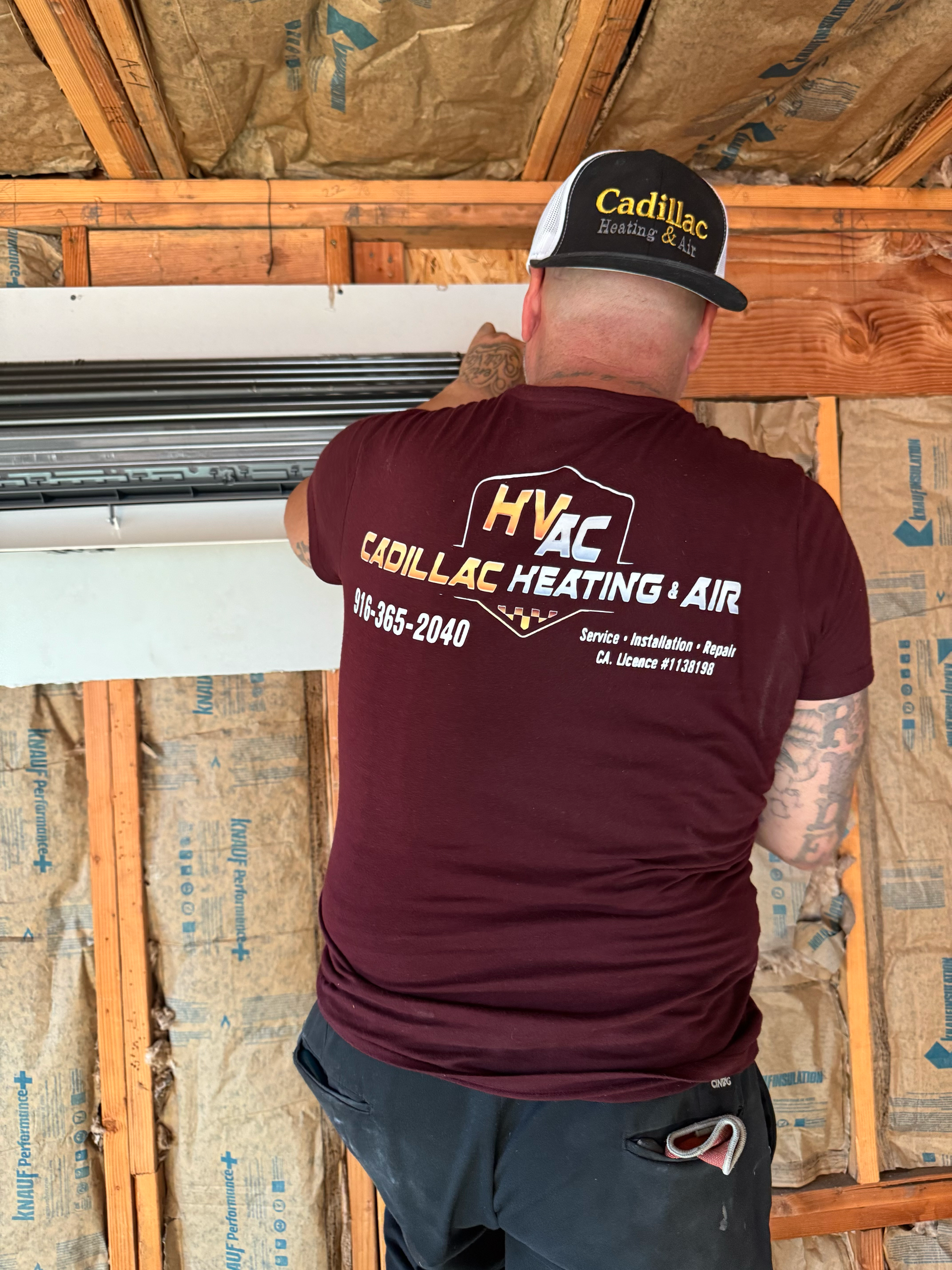 HVAC technician installs a unit, wearing a maroon shirt and a Cadillac hat, indoors with insulation.