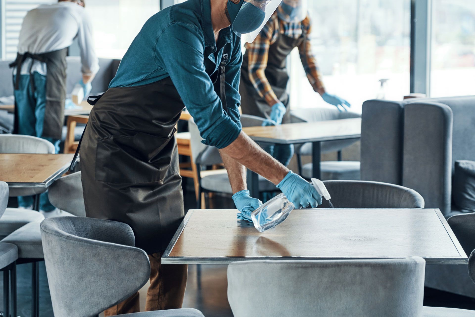 Staff in aprons, face shields, and gloves sanitizing tables in a restaurant to prevent the spread of germs.