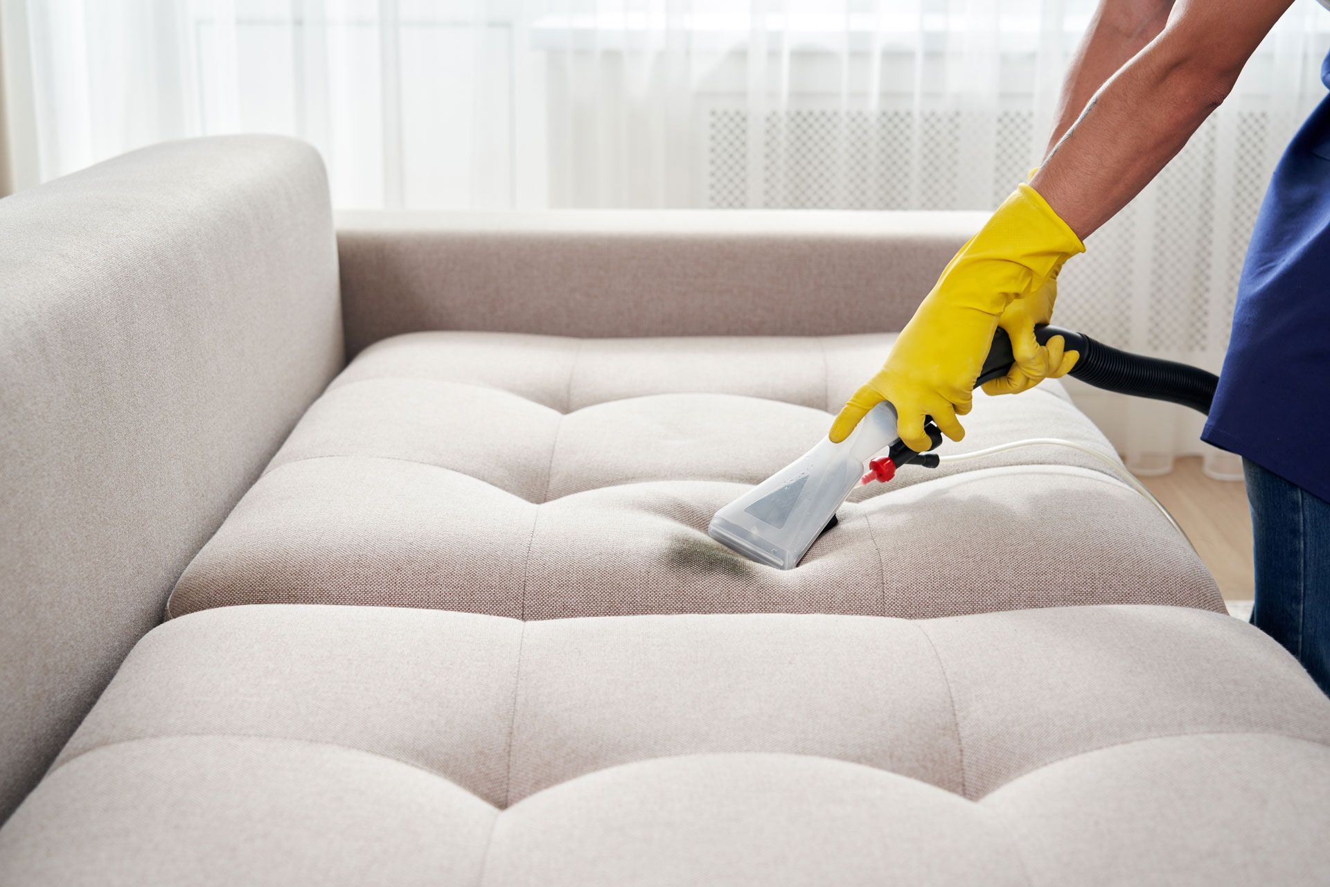 Person wearing yellow gloves using a cleaning machine on a light-colored couch in a well-lit room.