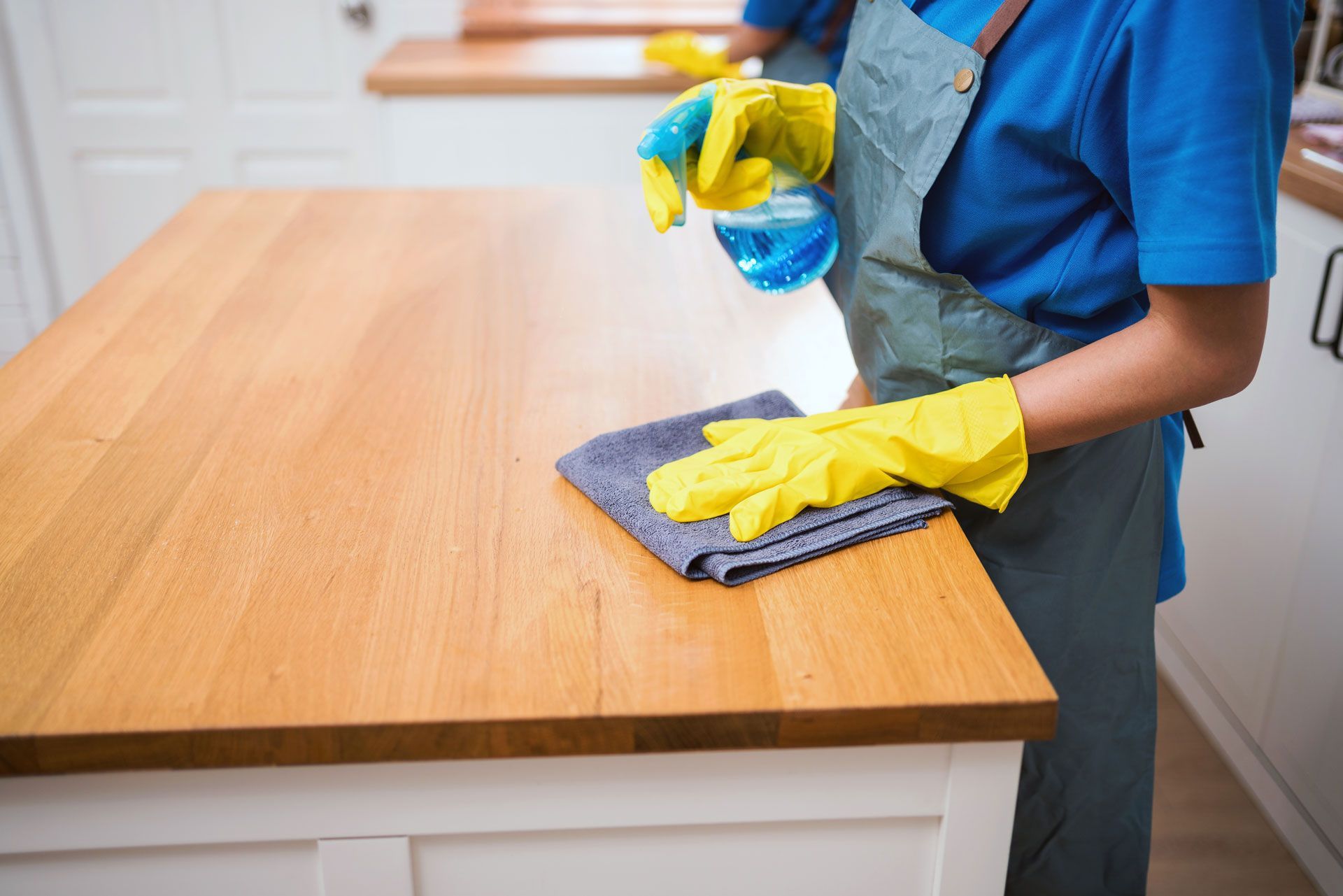 Person wearing blue apron and yellow gloves sprays and wipes a wooden countertop.
