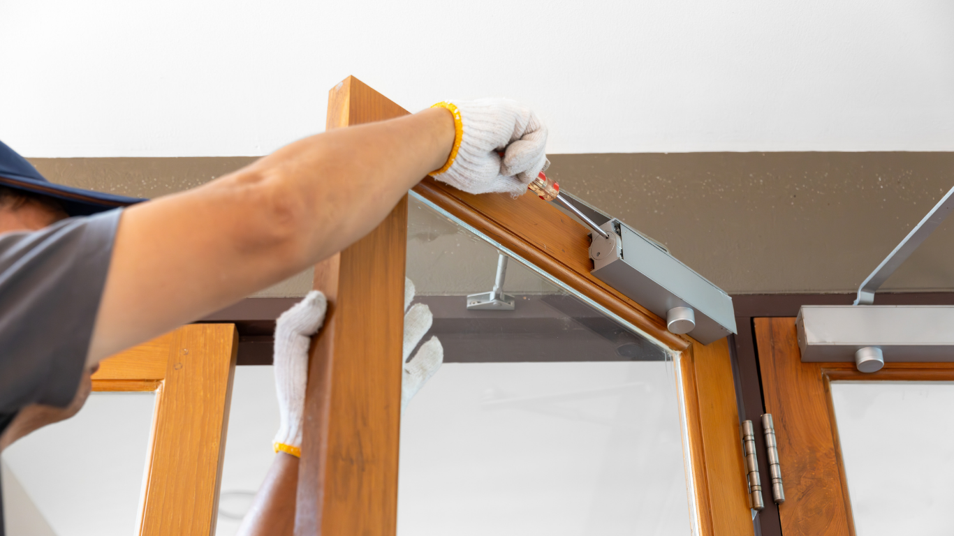 Person installing a door closer on a wooden door frame, wearing gloves.