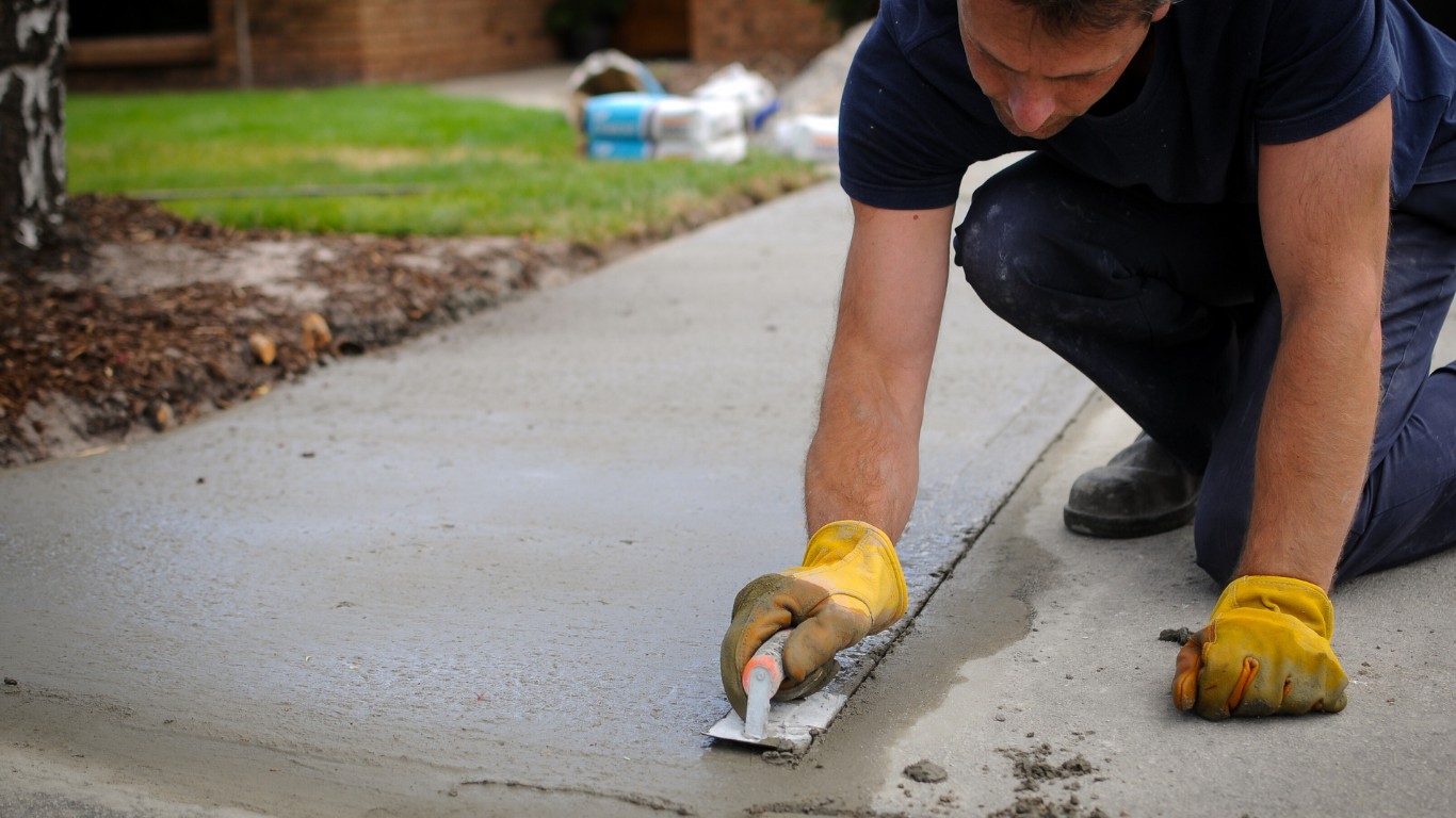 Man kneeling, smoothing cement crack on a sidewalk, wearing gloves, outdoors.