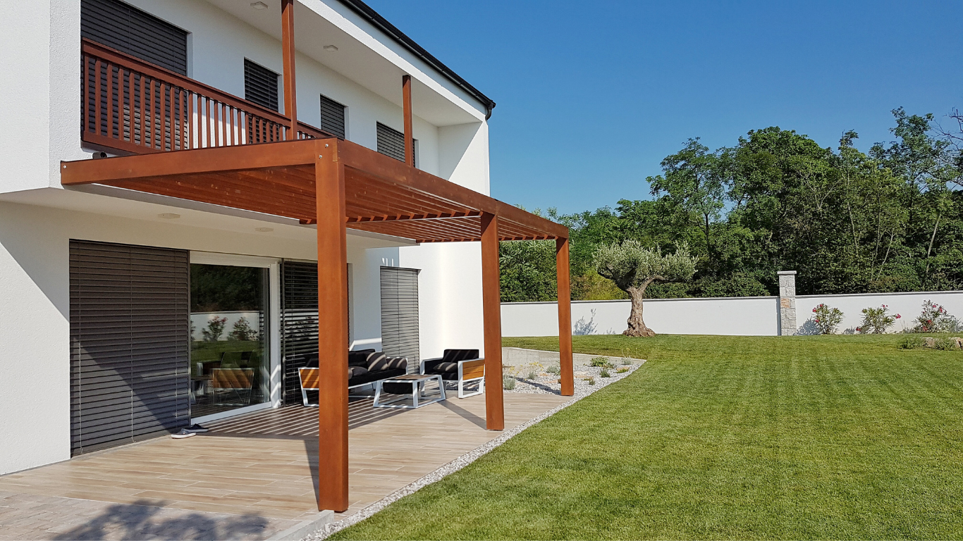A modern house with a wooden pergola over a patio, a grassy lawn, and trees under a blue sky.
