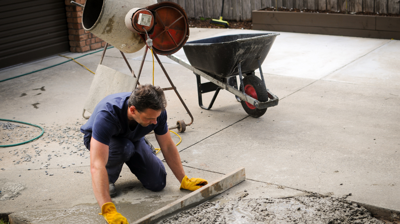 Man leveling fresh concrete with a tool, near a cement mixer and wheelbarrow on a driveway.