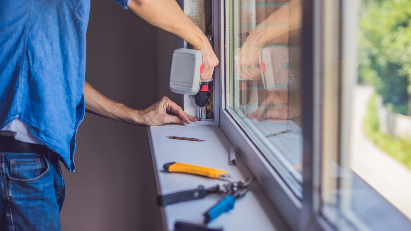 Person installing a window, using a power drill, on a windowsill. Blue shirt, jeans, sunny outdoors reflected.