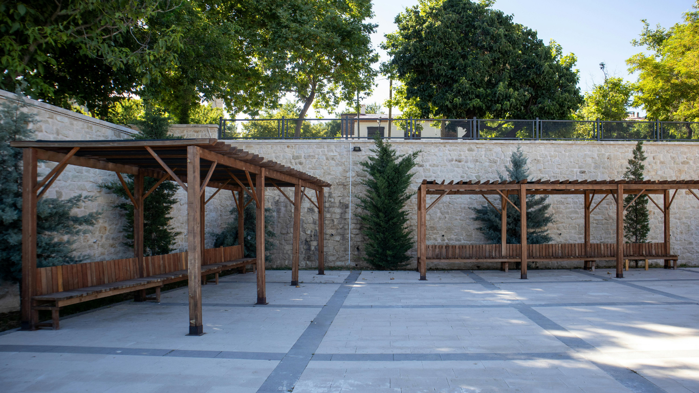 Two wooden pergolas with benches in a paved outdoor area, trees and a wall in background.