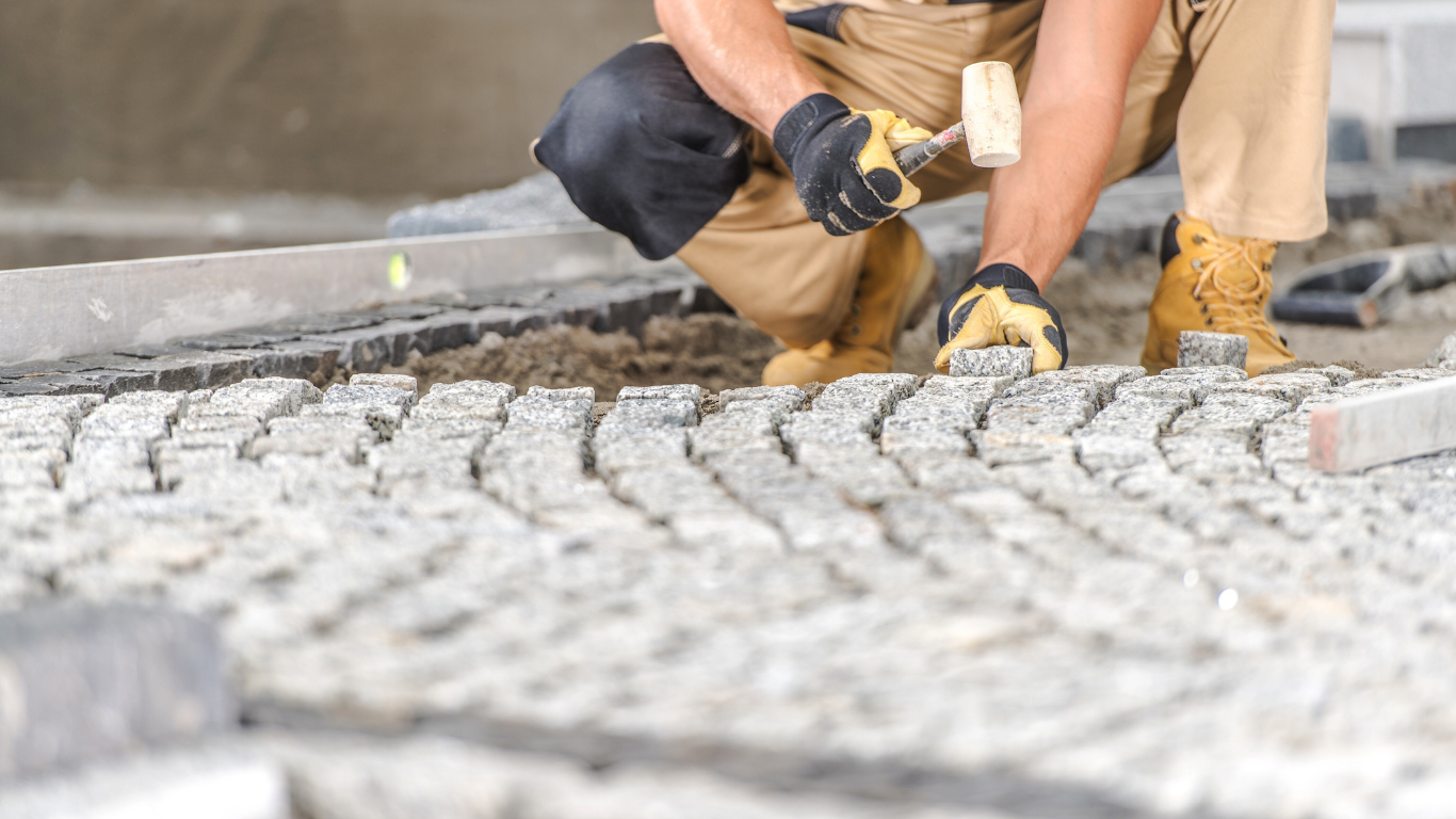 Construction worker kneeling, laying cobblestones with hammer and gloves.