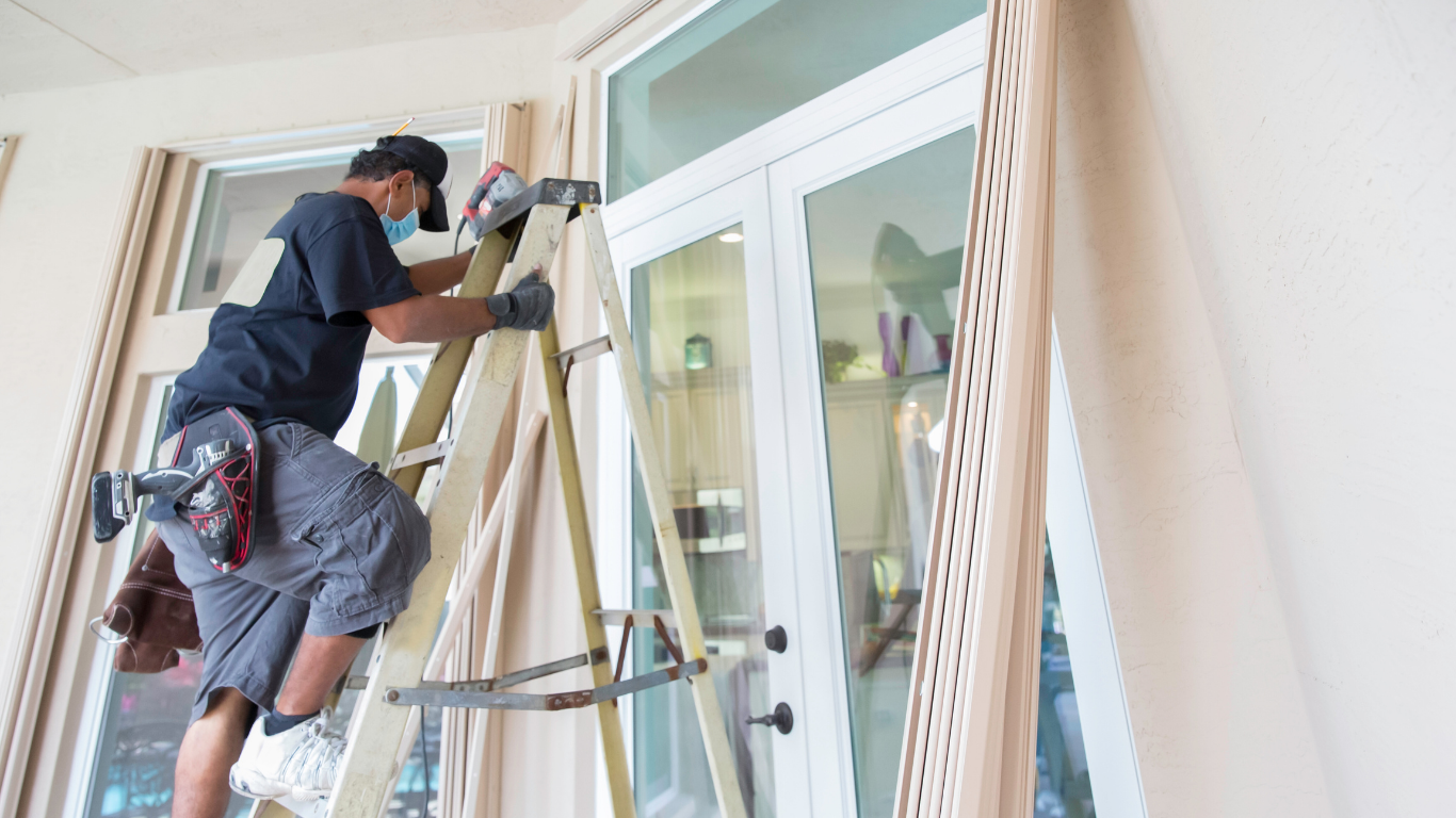 Man on ladder cleaning glass door in a bright room.
