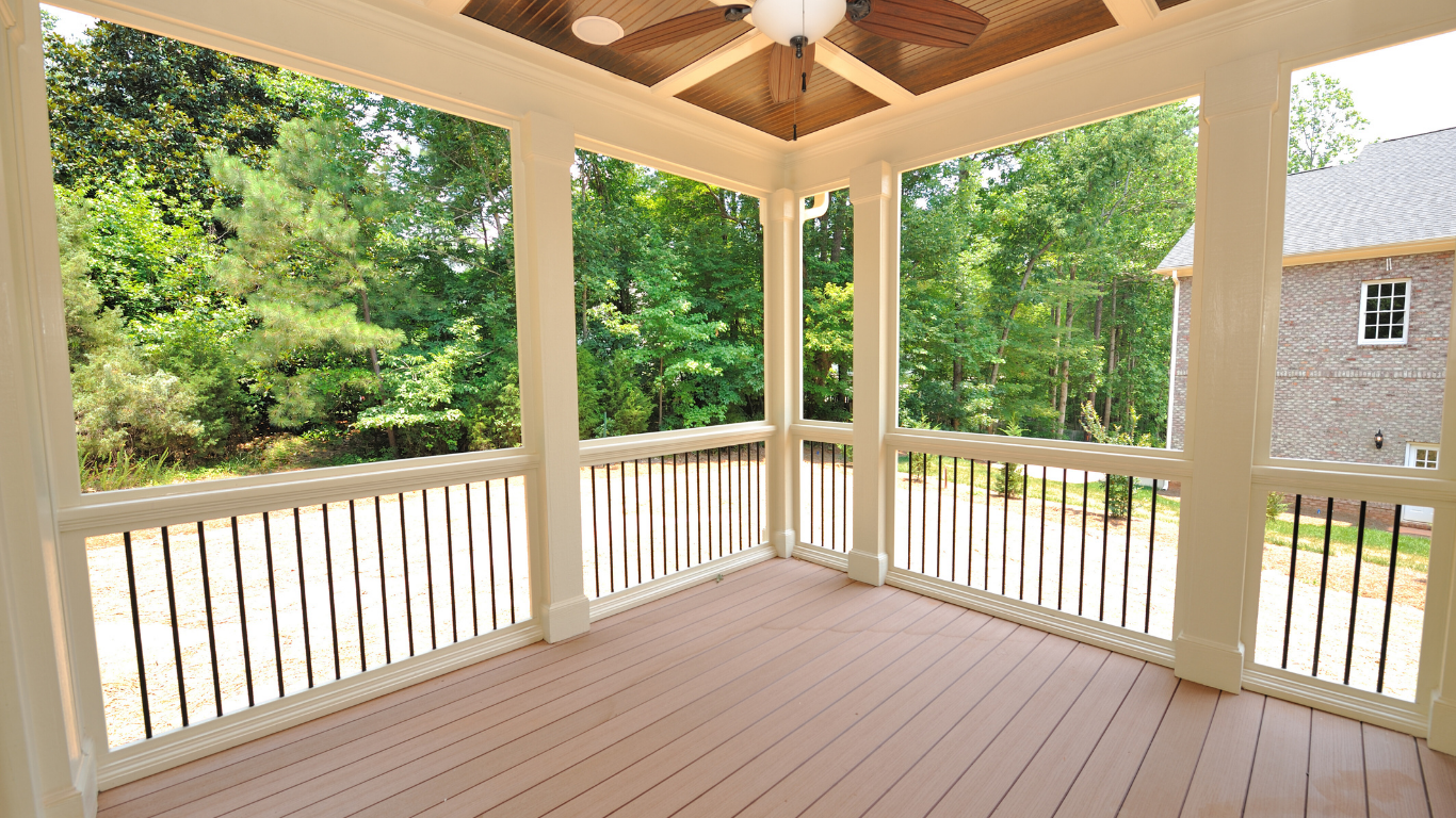 Screened porch with brown flooring and railings, white pillars, and a view of green trees.