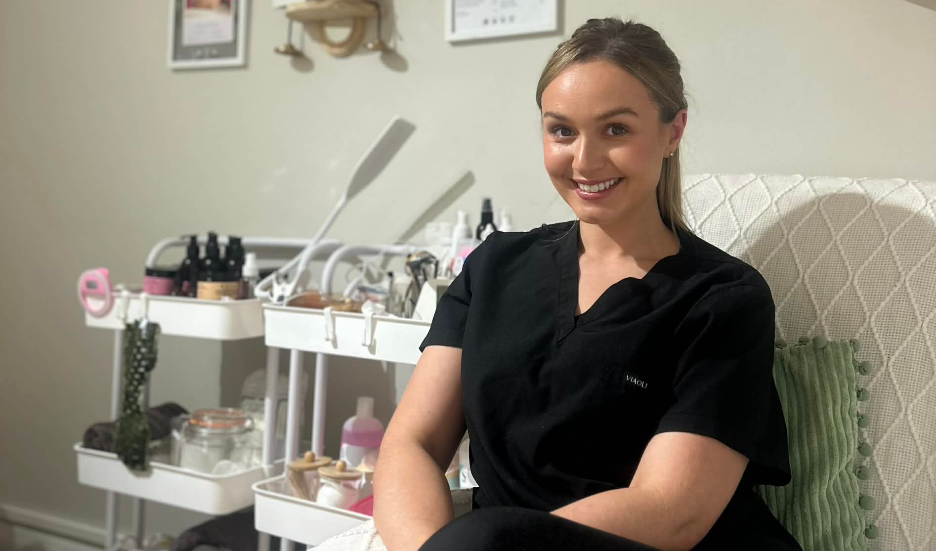 Smiling woman seated in a treatment room beside a manicure station and salon equipment