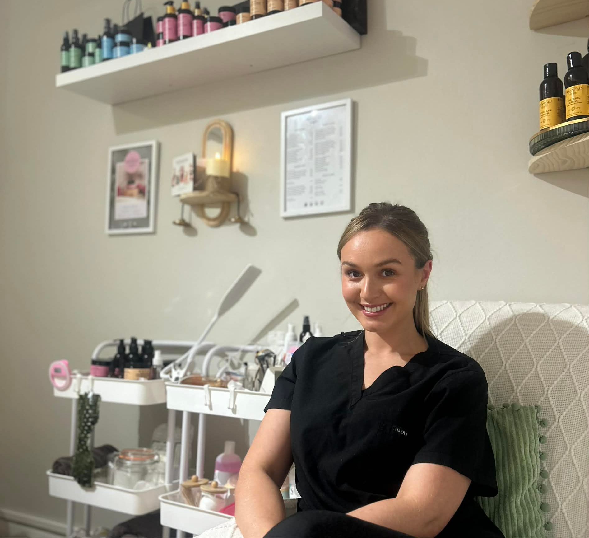 Woman smiling in a salon, seated beside nail products, shelves, and wall decor.