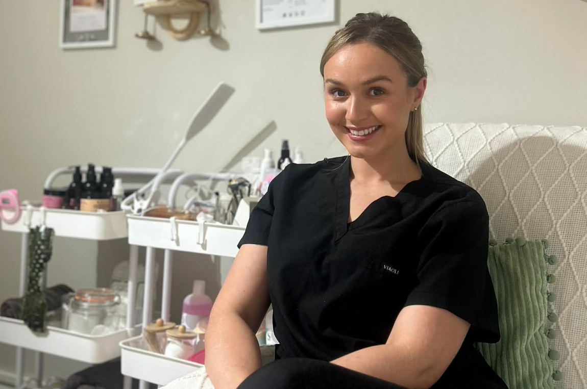 Woman smiling in black scrubs, seated in a bright beauty salon with manicure tools nearby
