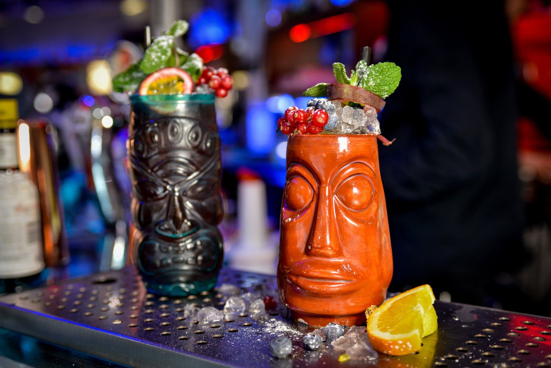 Due tazze tiki piene di cocktail, guarnite con frutta e menta, su un bancone.