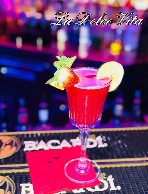 Un cocktail rosso vivace in un bicchiere con stelo, guarnito con una fragola, una fetta di limone e menta, servito su un bancone.