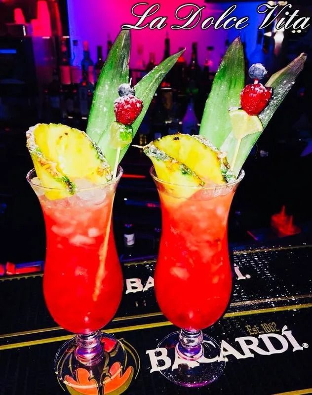 Due cocktail rossi in bicchieri alti, guarniti con ananas, lamponi e bordi zuccherati, su un bancone.