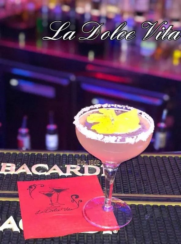 Cocktail servito in un bicchiere con bordo di zucchero, guarnizione di fiori gialli e tovagliolo rosso su un bancone con bottiglie sullo sfondo.