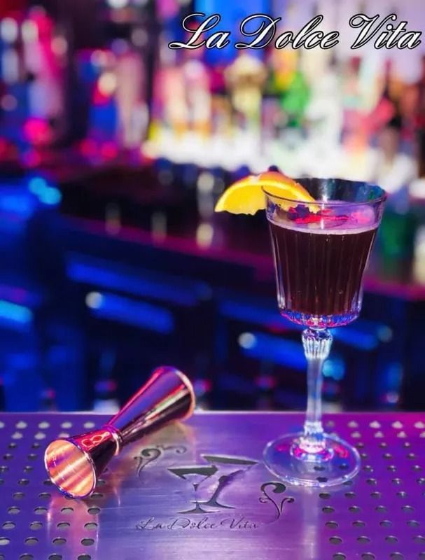 Cocktail scuro in un bicchiere elegante con guarnizione arancione; accanto a uno strumento di misurazione su un bancone del bar.