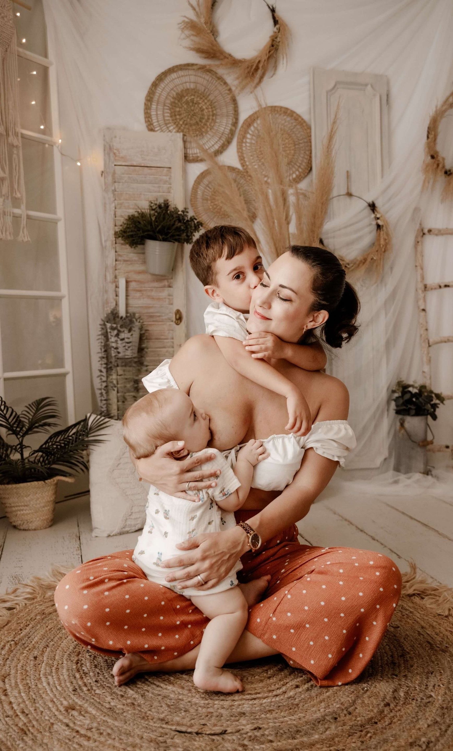 Una madre sentada sobre una alfombra tejida, sosteniendo a dos niños pequeños en una acogedora habitación de estilo bohemio con decoración neutra.