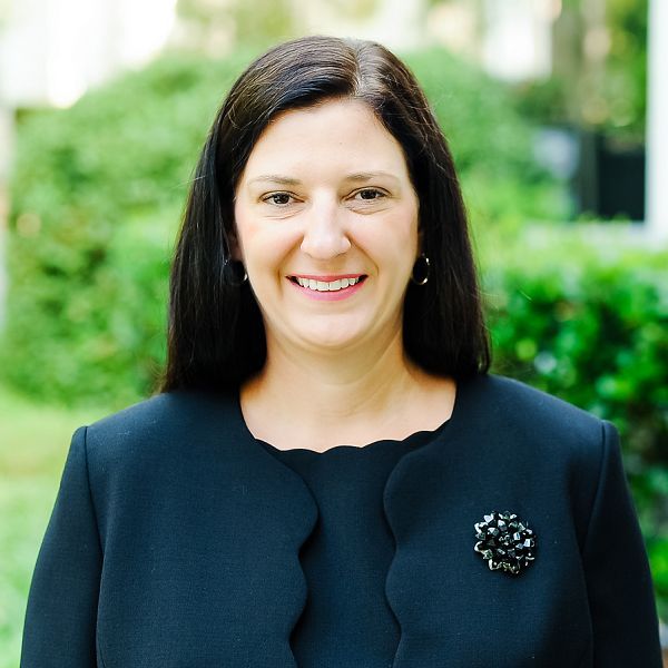 A woman wearing a black jacket and a brooch smiles for the camera