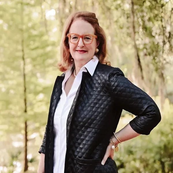 A woman wearing glasses and a black jacket is standing in the woods.