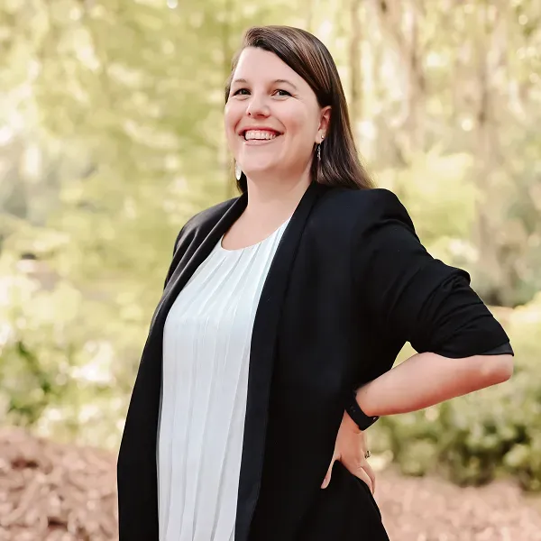 A woman in a black jacket and white shirt is smiling while standing in the woods.