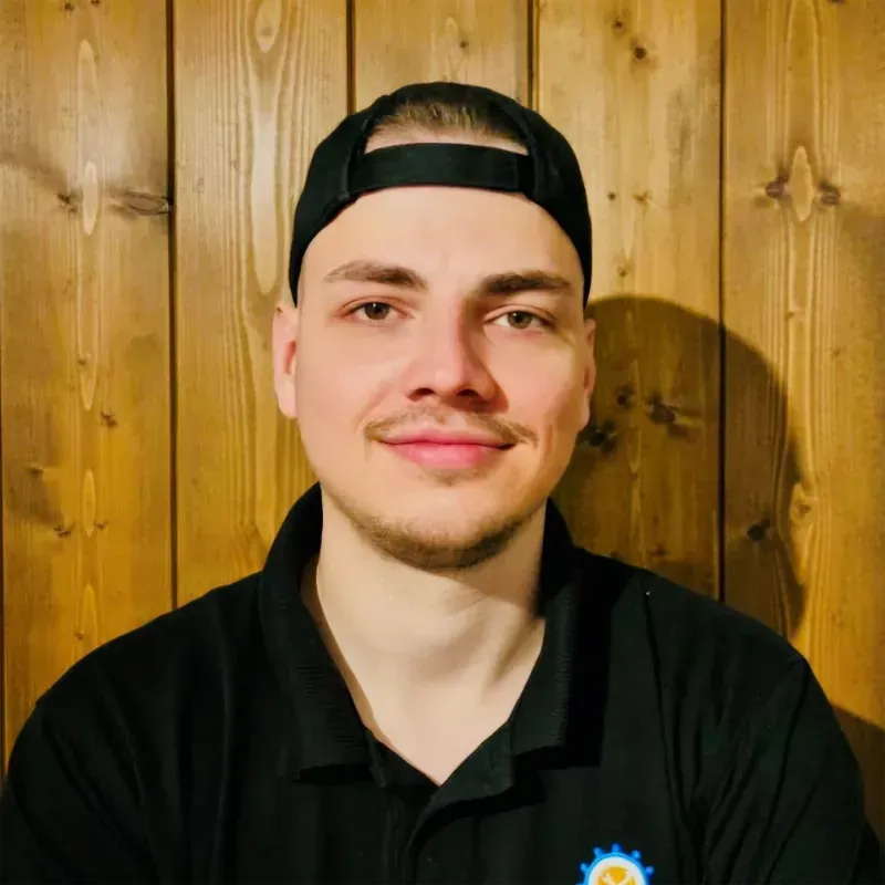 Ein Mann mit schwarzer Kappe und schwarzem Hemd lächelt in die Kamera, vor einer Holzvertäfelung stehend.