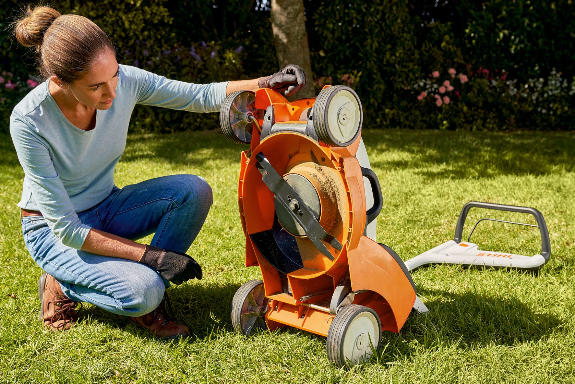 A woman is kneeling down next to a lawn mower on the grass.