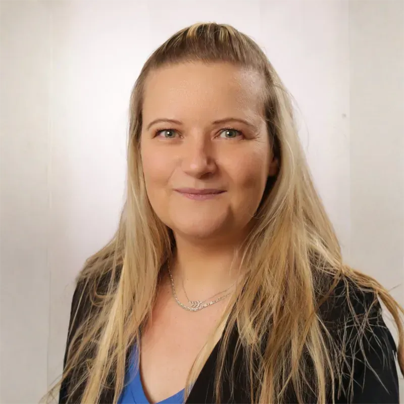 Eine blonde Frau, die eine schwarze Jacke und ein blaues Hemd trägt, lächelt und trägt eine silberne Halskette.