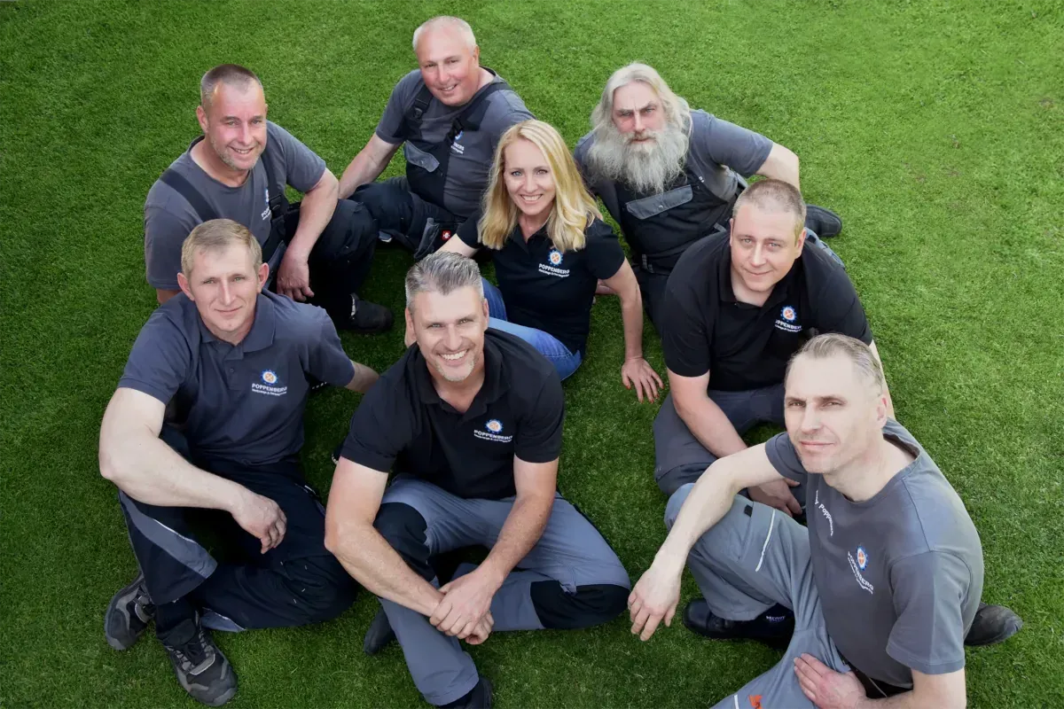 Eine Gruppe von acht Personen in Arbeitskleidung (Mitarbeiter der Firma POPPENBERG aus Altlandsberg) sitzt zusammen auf einer Rasenfläche und lächelt in die Kamera.