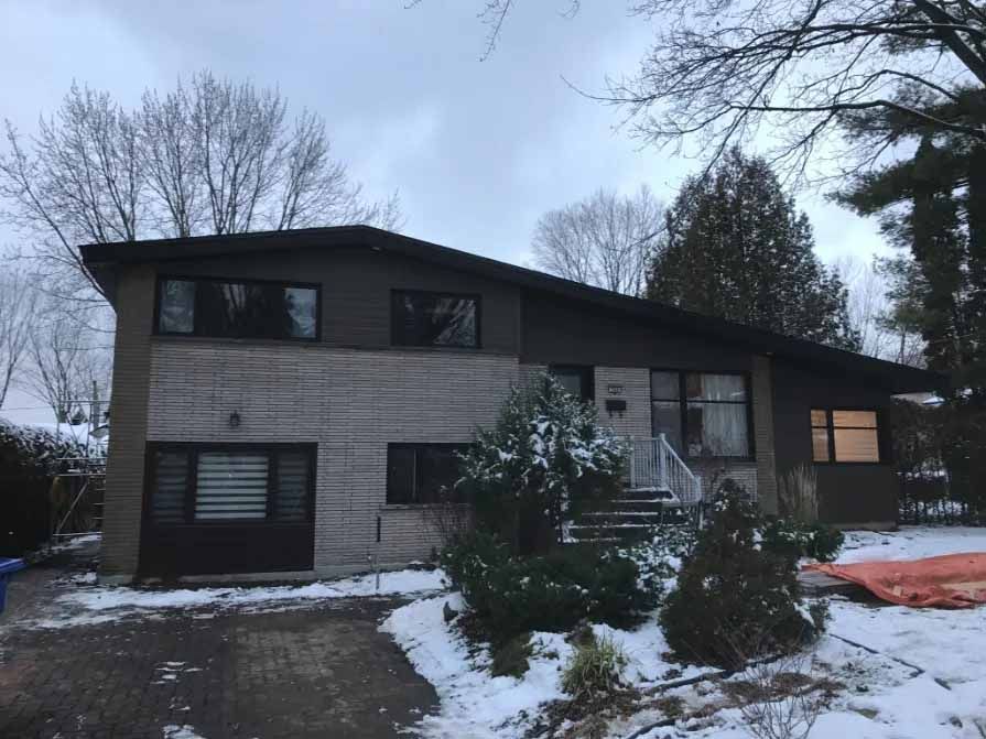 Maison à deux étages avec façade en briques grises et boiseries brun foncé, neige au sol, journée nuageuse.