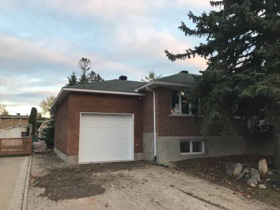 Garage en construction, vue extérieure. Bâche bleue sur les murs, porte de garage ouverte, bois et allée visibles. Ciel nuageux.